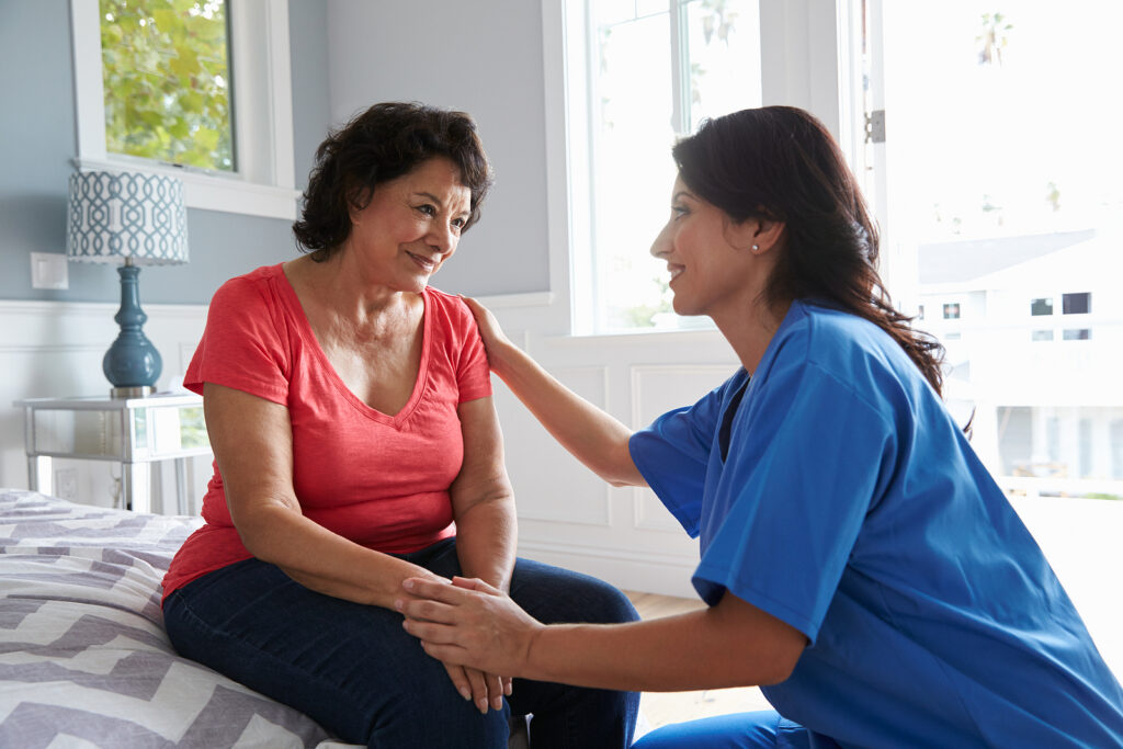 Caregiver in blue scrubs sits beside a woman in a red shirt on a bed, offering comfort with a hand on her shoulder.